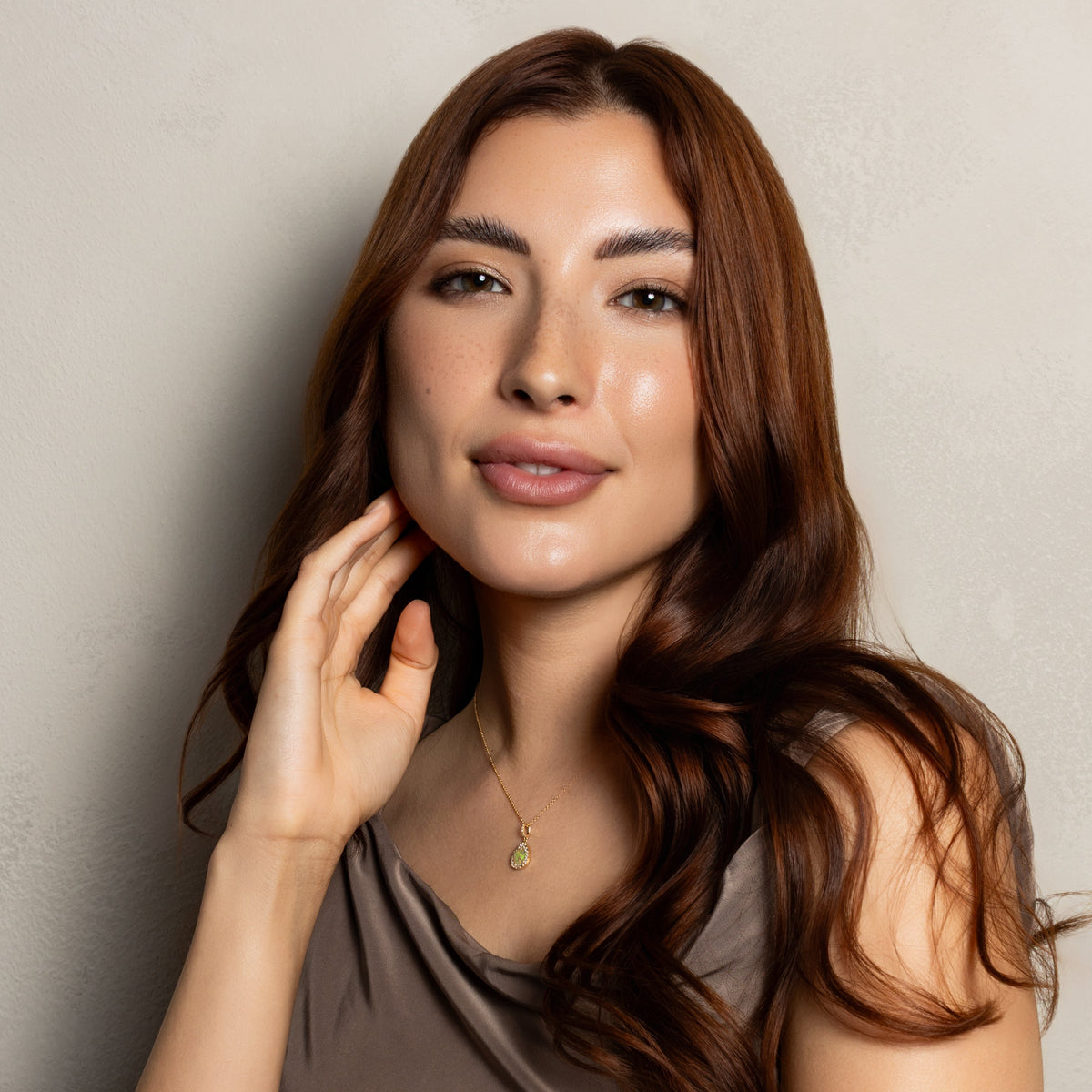 A woman with long, wavy brown hair is shown from the chest up against a plain light gray wall. She is wearing a sleeveless taupe-brown top and a delicate gold necklace with a small green pendant.