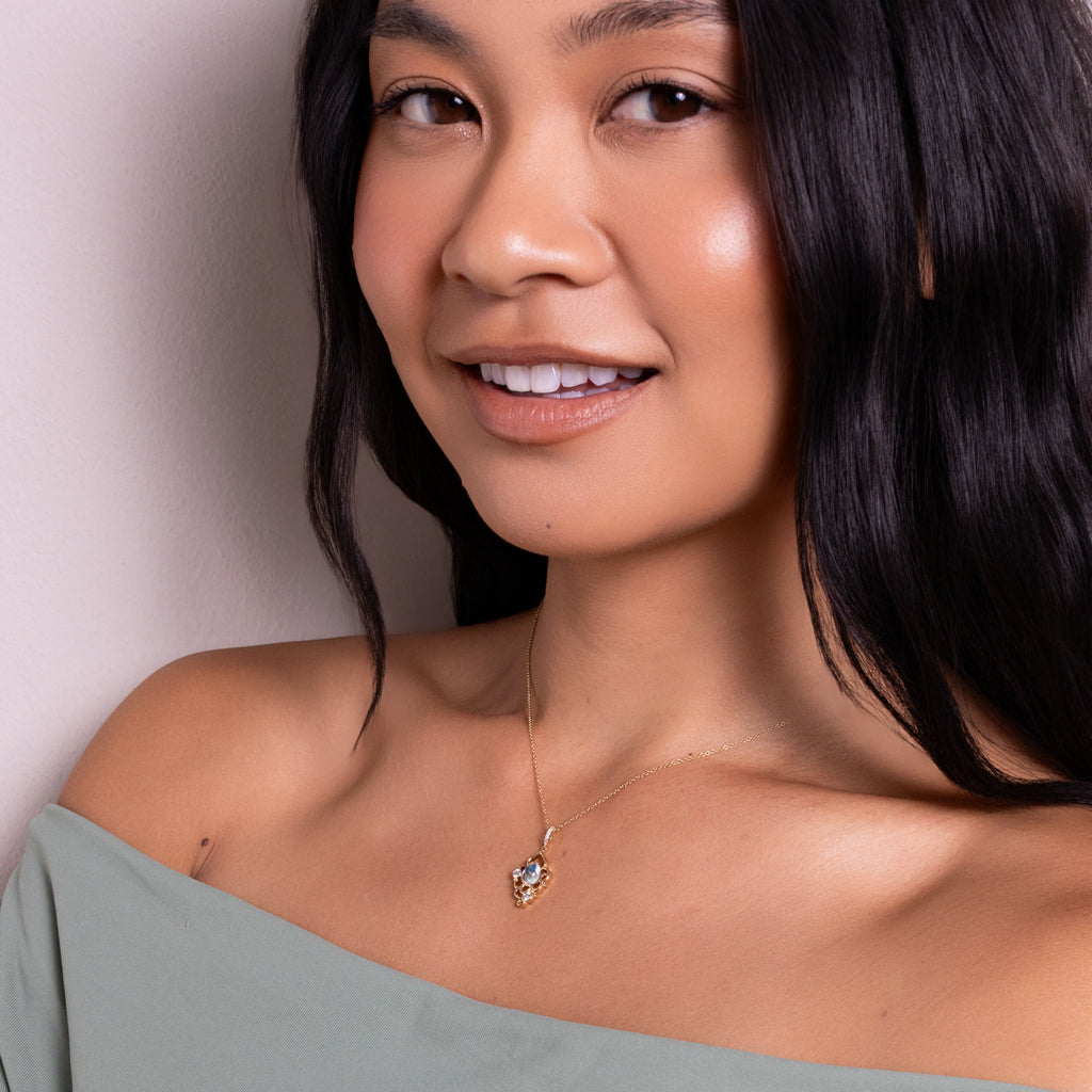 A close-up lifestyle shot of a woman wearing a delicate gold necklace with a small heart-shaped crystal pendant, paired with an off-the-shoulder sage green top.