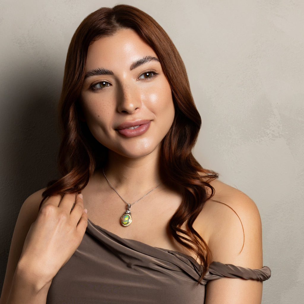 A woman with long wavy brown hair wearing a taupe one-shoulder top and a silver pendant necklace with a green stone, posing against a textured gray wall.