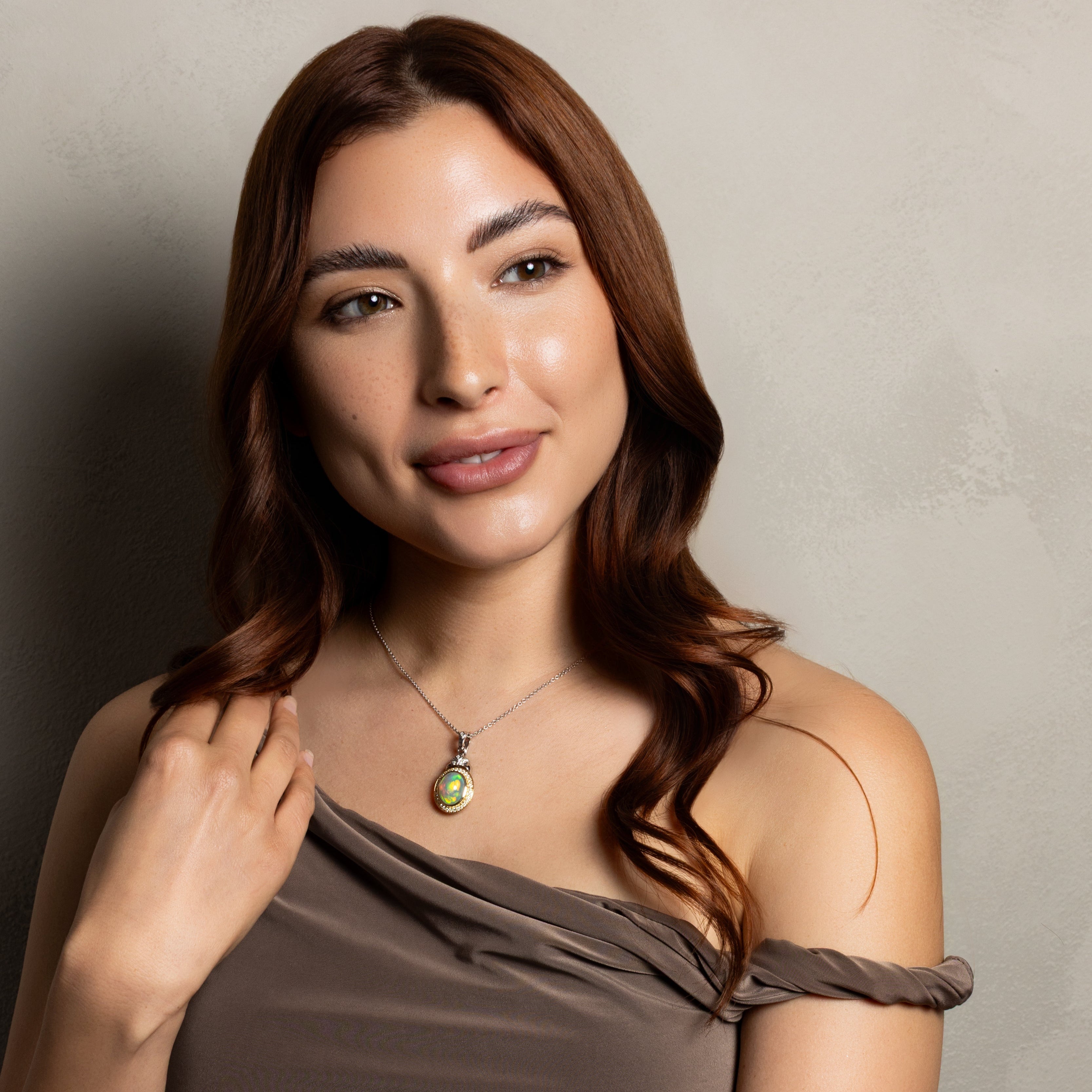A woman with long wavy brown hair wearing a taupe one-shoulder top and a silver pendant necklace with a green stone, posing against a textured gray wall.