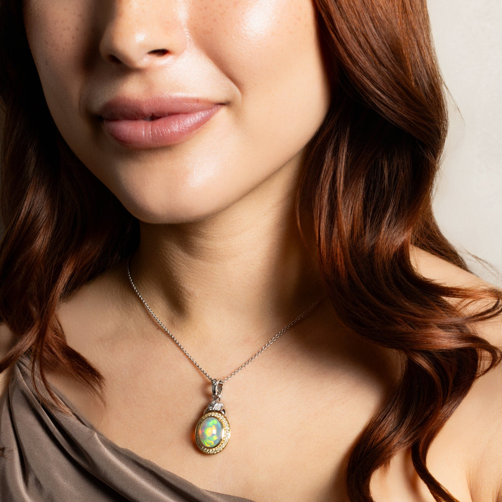 A close-up of a woman wearing a silver-toned pendant necklace with an oval iridescent green and gold cabochon stone, paired with a taupe draped top.