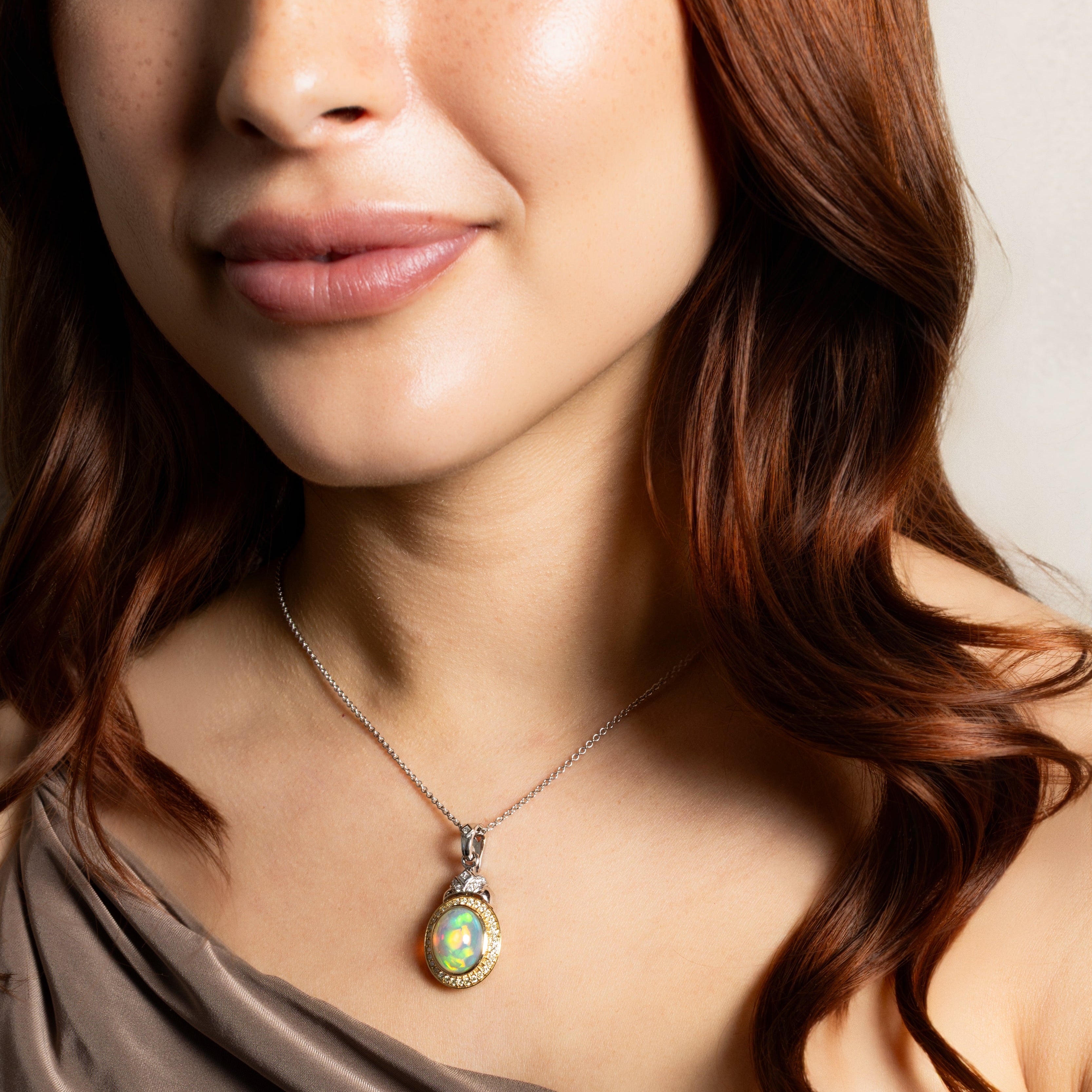 A close-up of a woman wearing a silver-toned pendant necklace with an oval iridescent green and gold cabochon stone, paired with a taupe draped top.