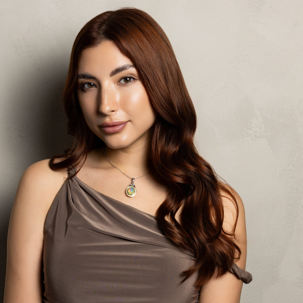 A woman with long, wavy brown hair wearing a draped, one-shoulder taupe-brown dress and a small pendant necklace, posing against a textured light gray wall.