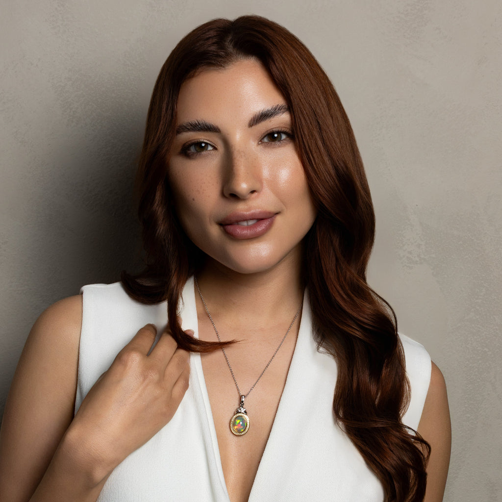 A woman with long brown hair wearing a white sleeveless wrap top and a gold pendant necklace with a green and gold circular charm, posing against a textured gray wall.