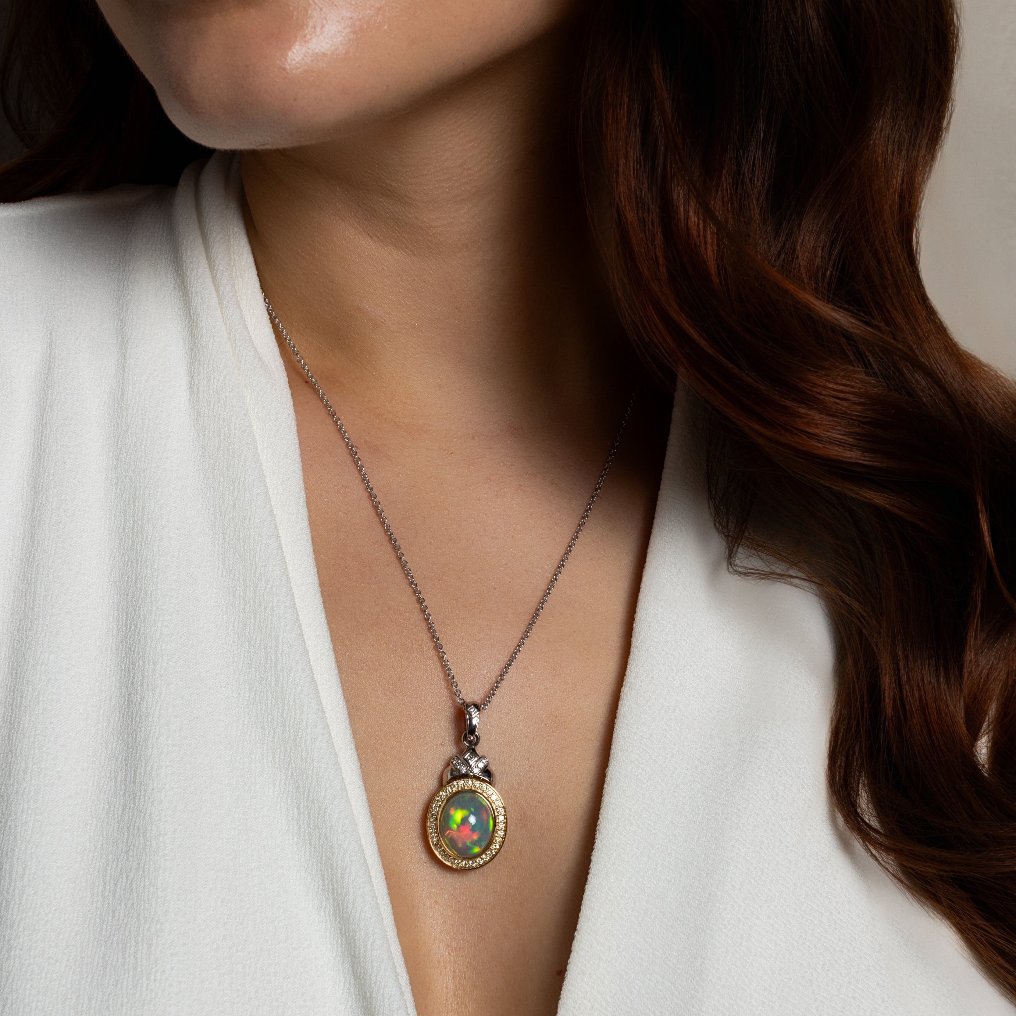 A close-up of a woman wearing a silver pendant necklace with a colorful, iridescent oval inlay. The necklace is shown against her white blouse and brown hair.