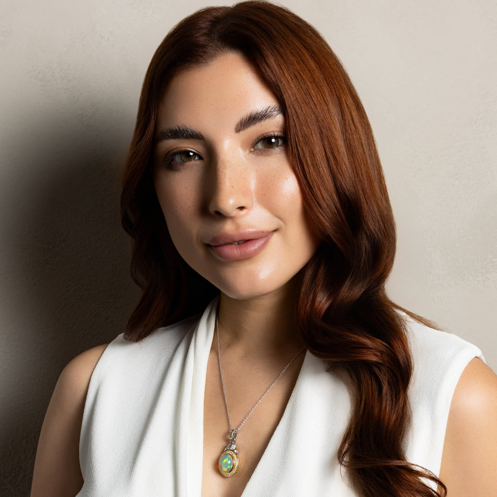 A woman with long, wavy auburn hair is shown from the chest up, wearing a white sleeveless wrap-style top and a silver pendant necklace with a green and gold oval stone. She is posing against a neutral, warm-toned textured wall.
