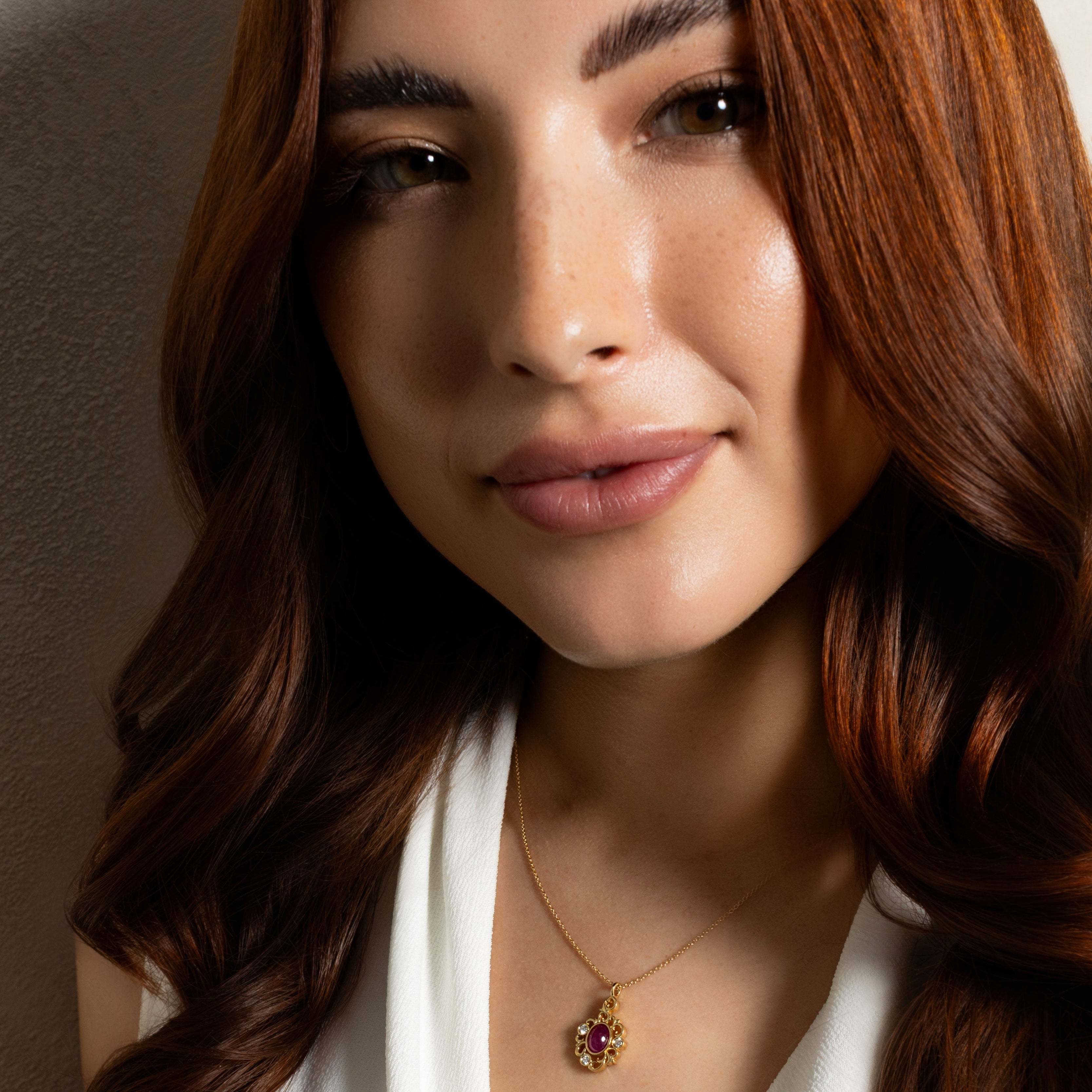 A close-up of a woman with long wavy brown hair wearing a delicate gold necklace with a small round pendant featuring a red and yellow stone, paired with a white V-neck top.