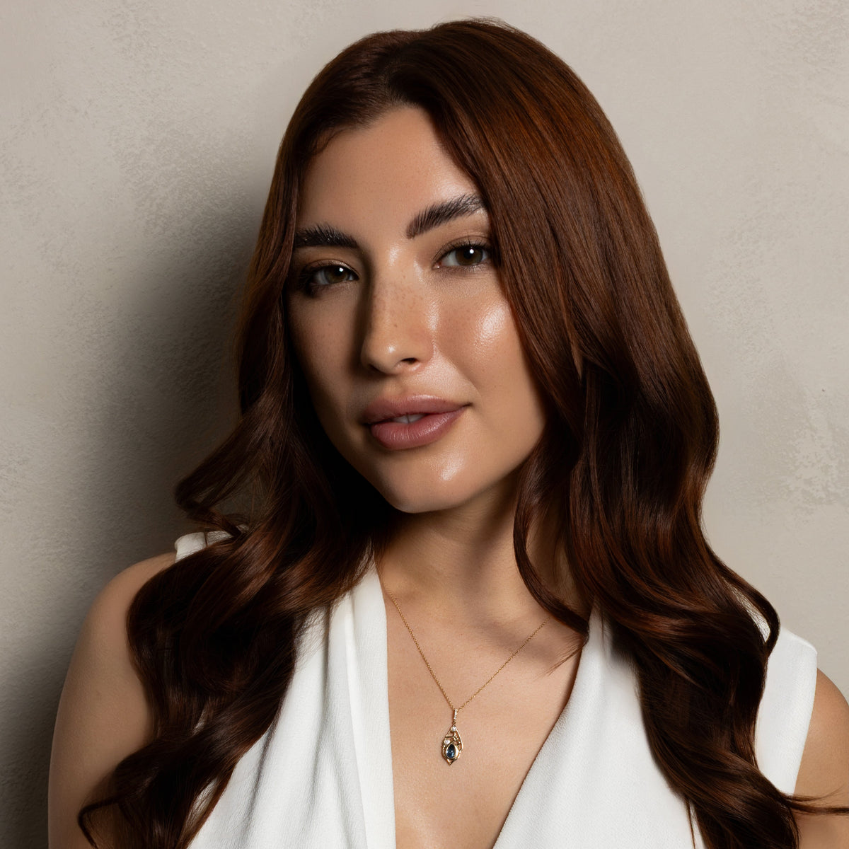 A woman with long, wavy brown hair wearing a white sleeveless wrap top and a delicate gold necklace with a small blue stone pendant, posing against a neutral textured beige wall.