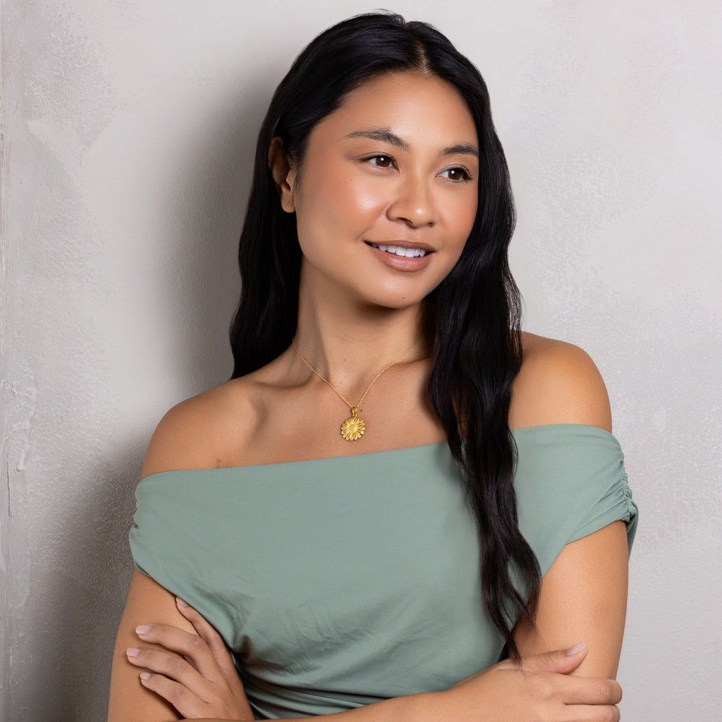 A woman with long black hair wearing an off-the-shoulder sage green top and a gold pendant necklace, posing with her arms crossed against a textured light gray wall.