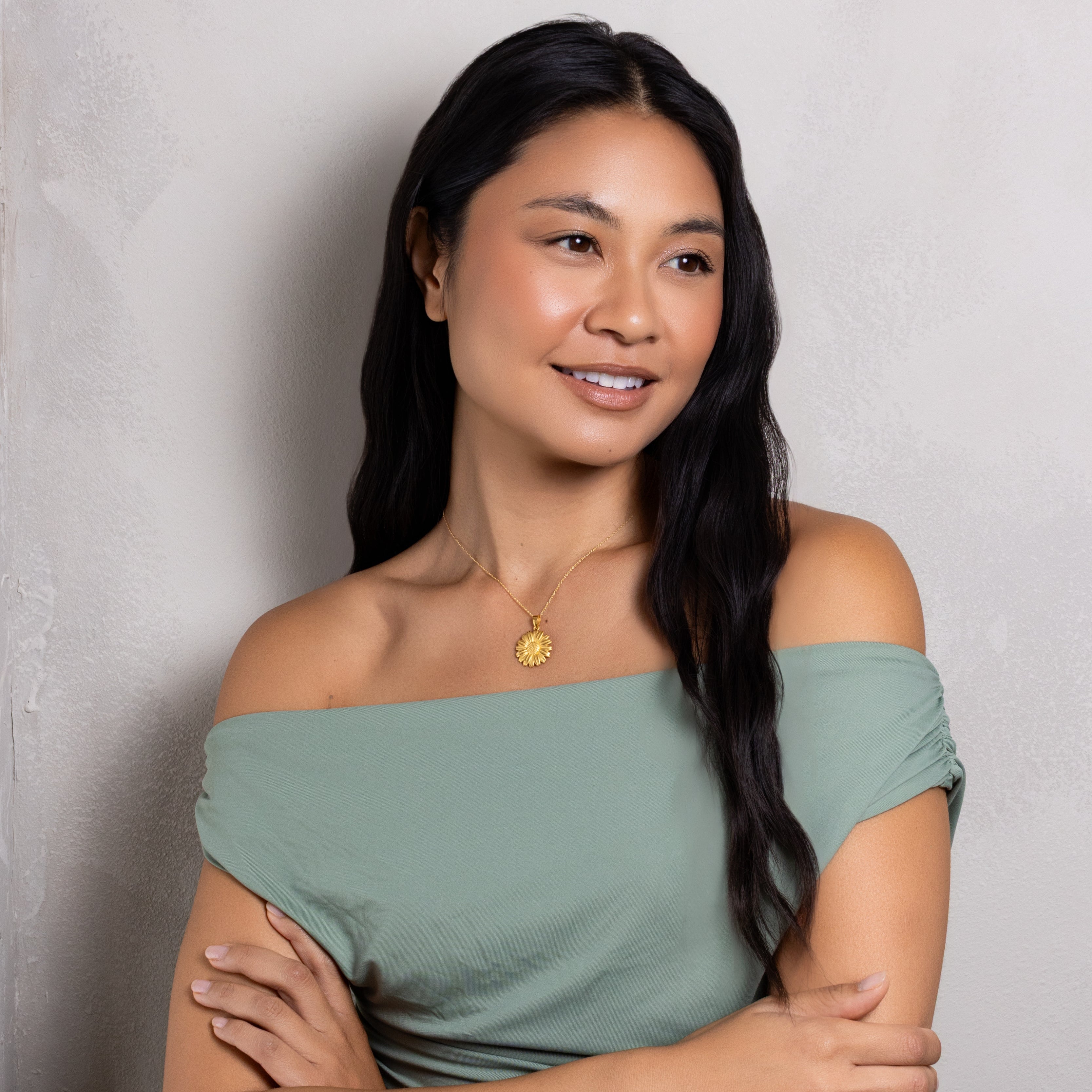 A woman with long black hair wearing an off-the-shoulder sage green top and a gold pendant necklace, posing with her arms crossed against a textured light gray wall.