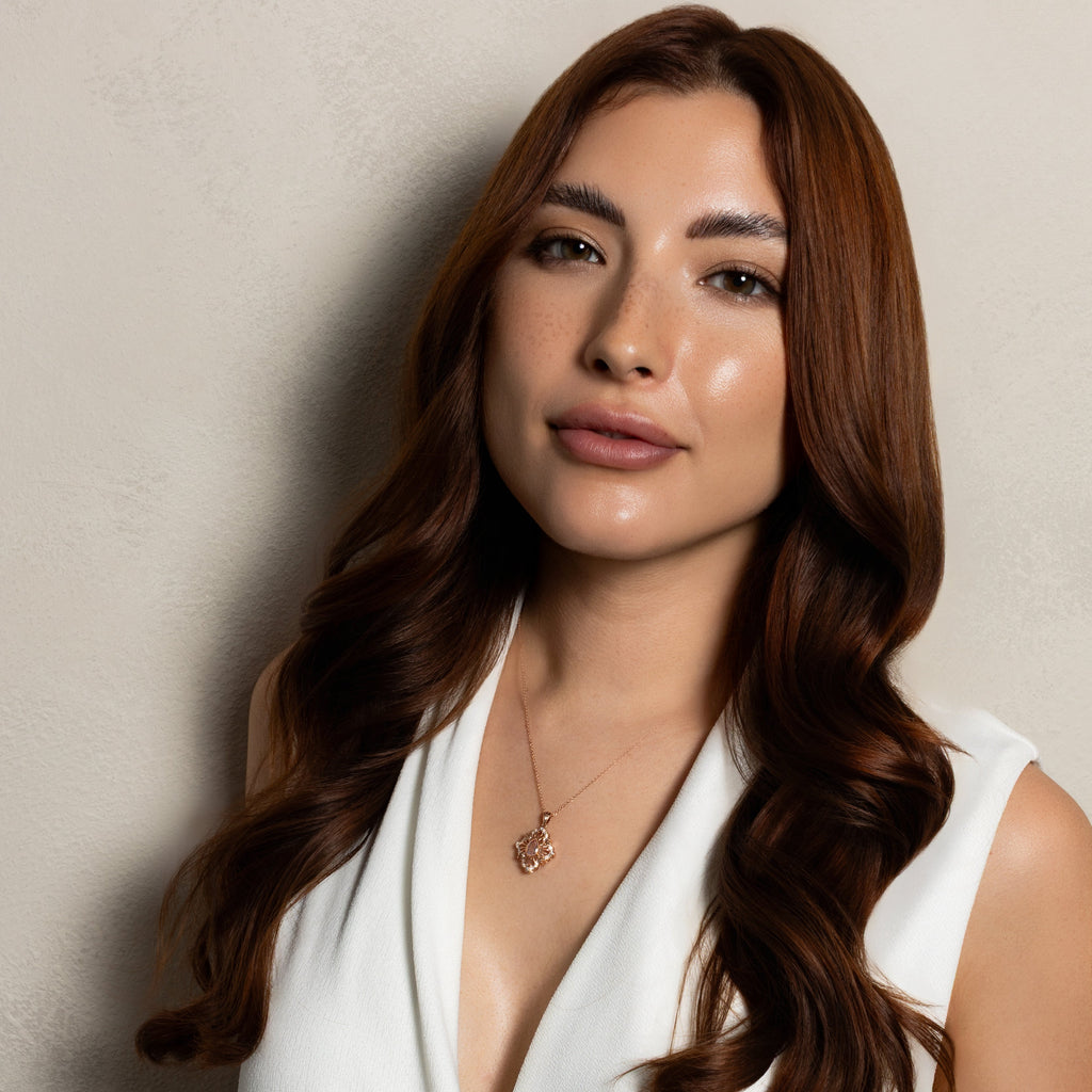 A woman with long, wavy brown hair wearing a white sleeveless wrap-style top, photographed against a plain light beige wall.