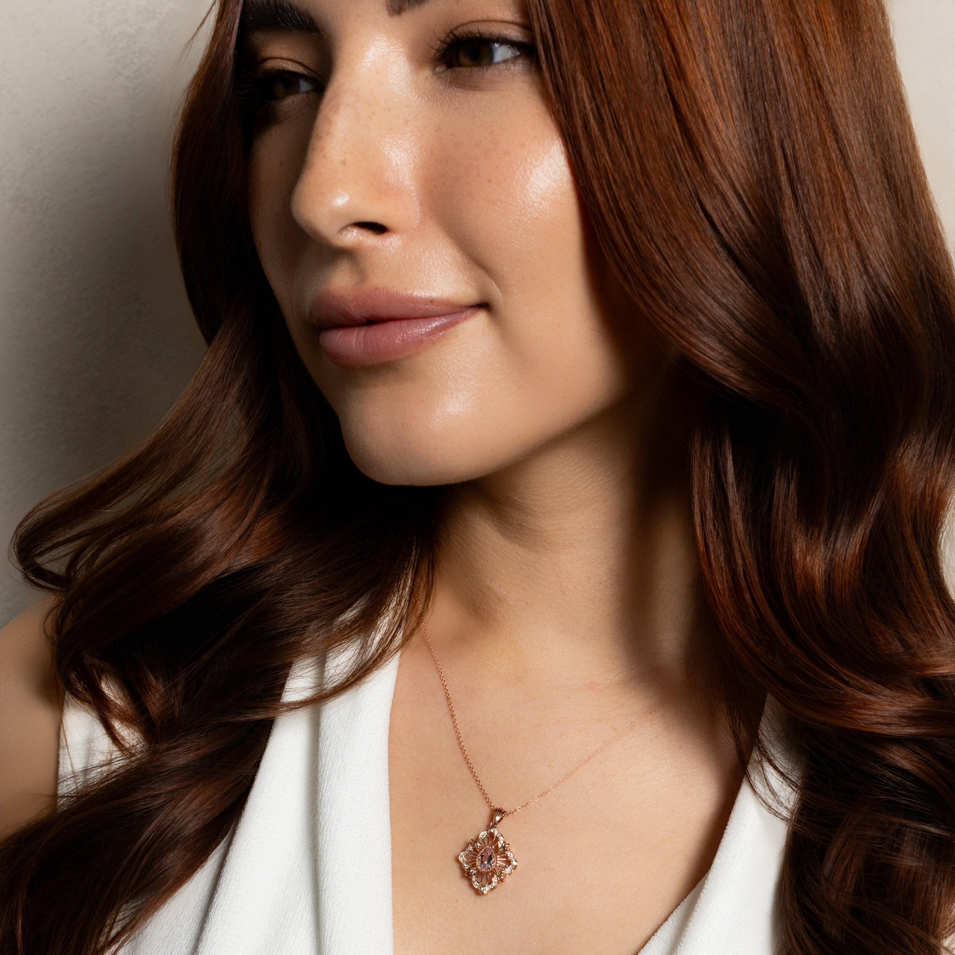 A close-up lifestyle shot of a woman with long, wavy brown hair wearing a delicate rose gold pendant necklace and a white V-neck top.