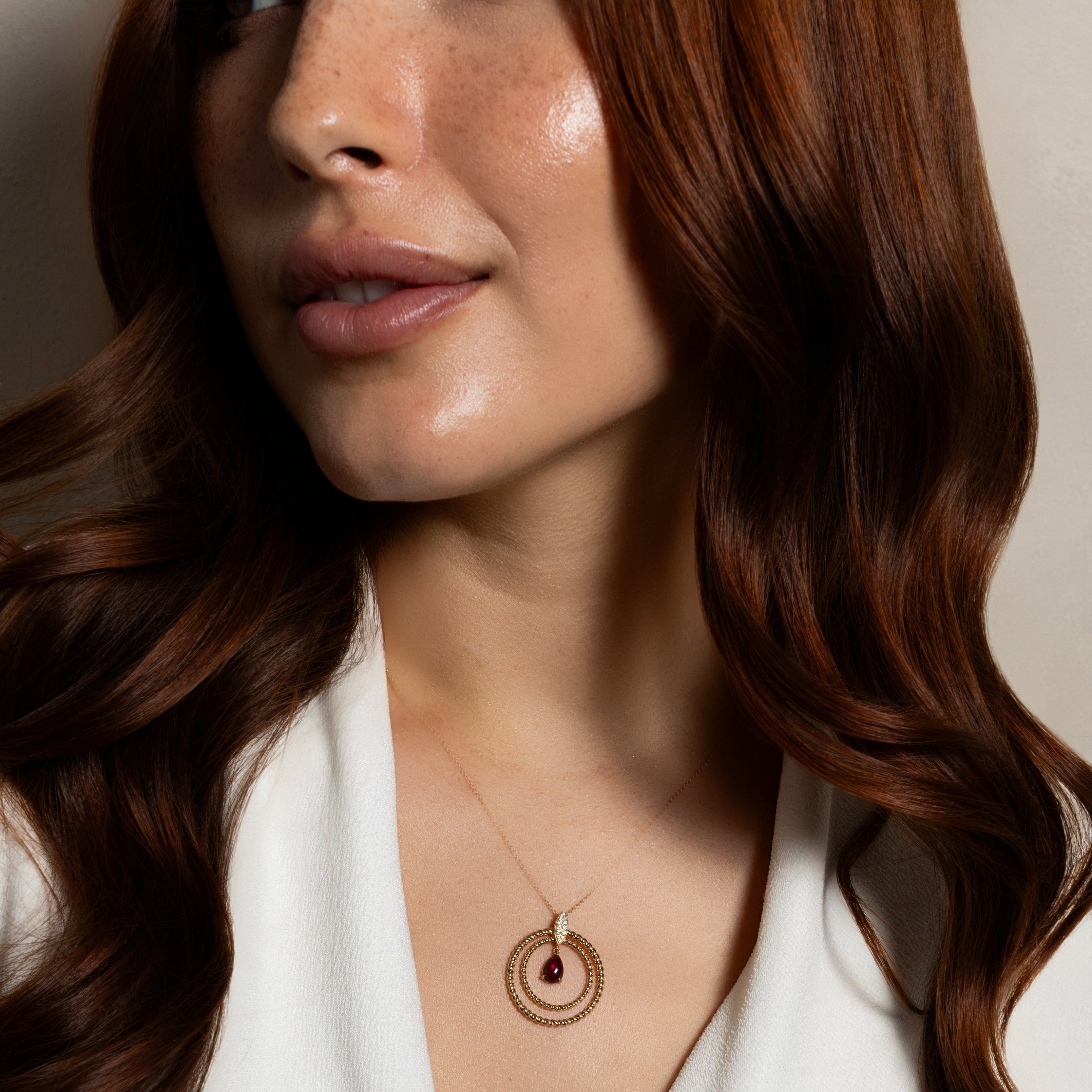 A close-up of a woman with long wavy brown hair wearing a delicate gold necklace with a double-ring pendant and a small dark teardrop gemstone. She is wearing a white V-neck top against a soft beige background.