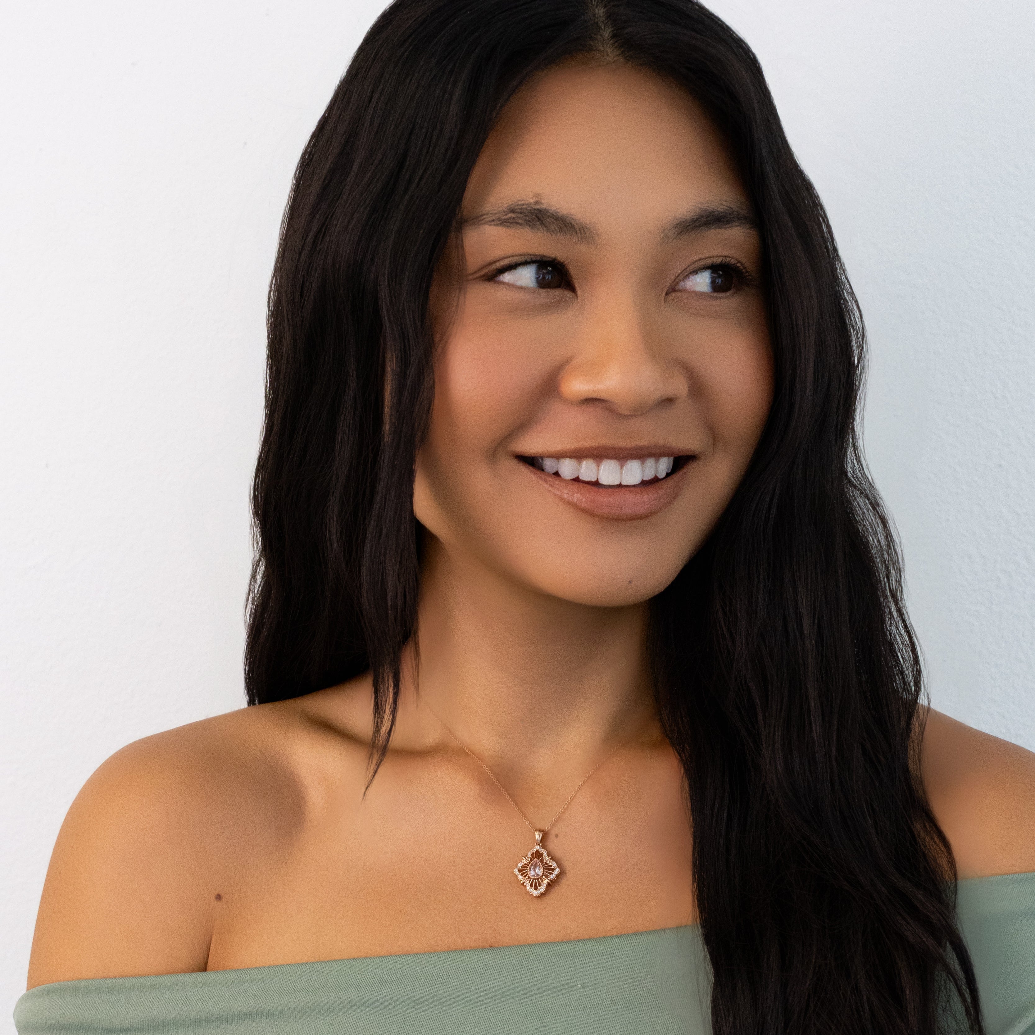 A woman with long dark hair wearing an off-the-shoulder sage green top and a delicate gold pendant necklace, photographed against a plain light gray background.