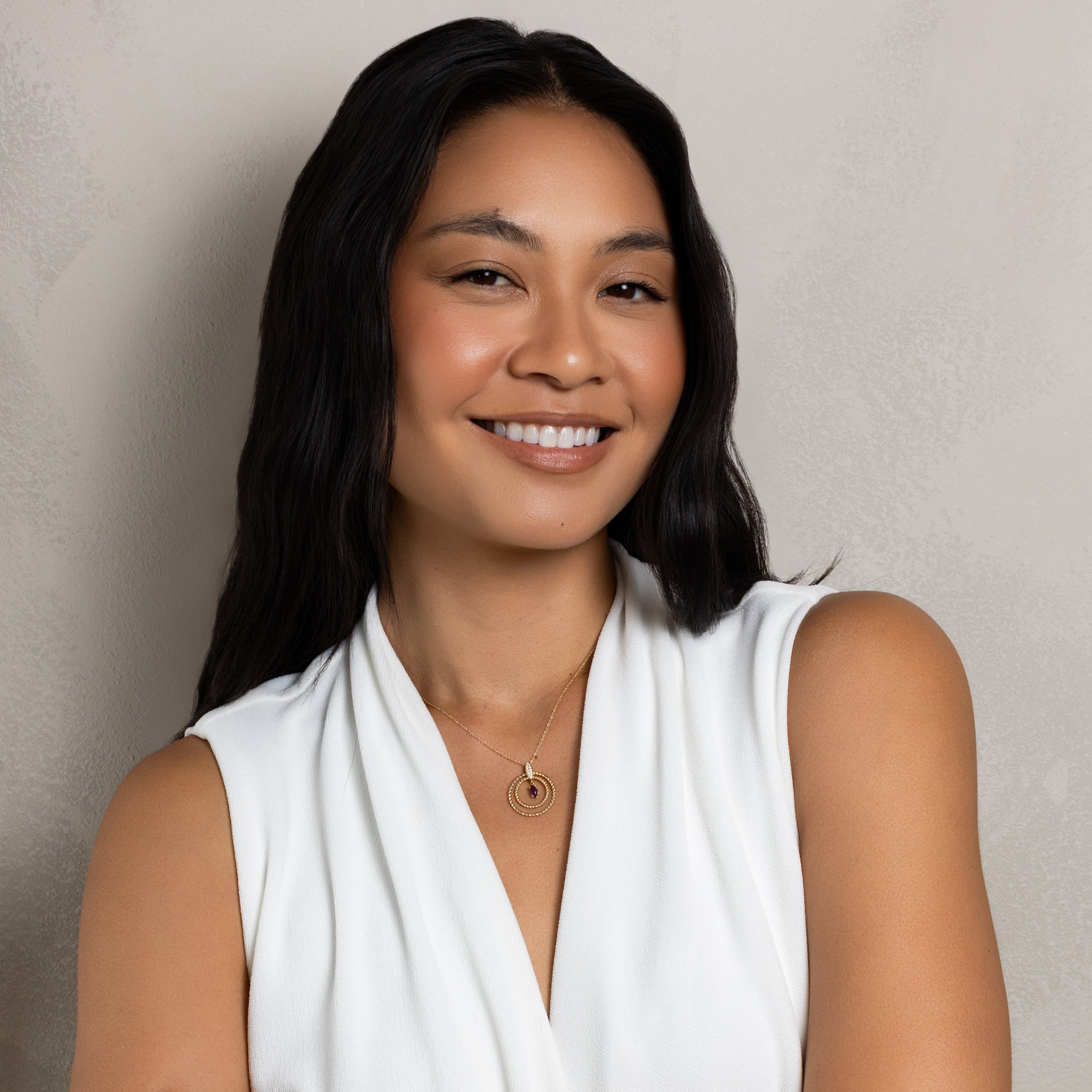 A smiling woman with long dark hair wearing a white sleeveless draped top, standing with her arms crossed against a textured light gray wall.