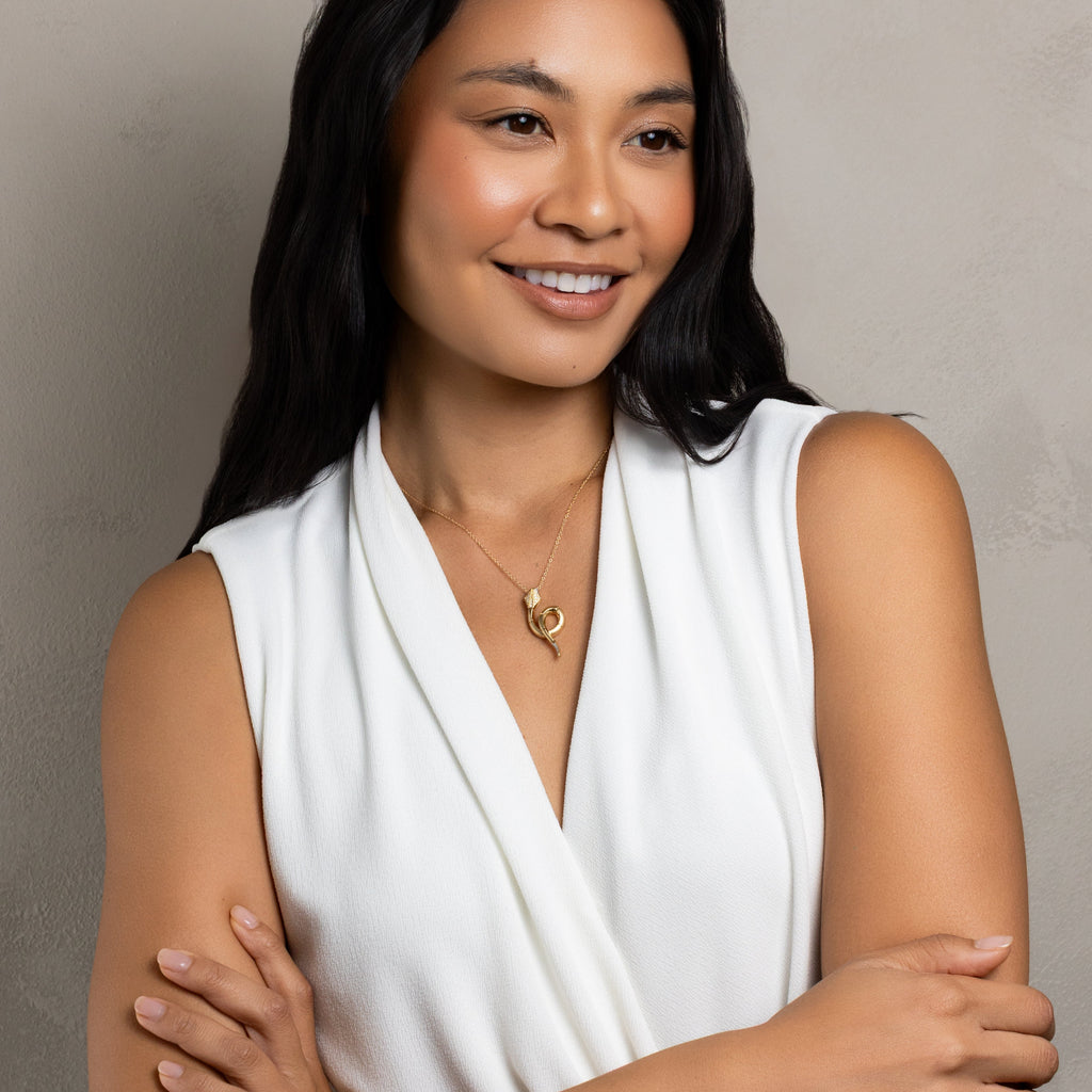 A smiling woman with long dark hair wearing a white sleeveless draped top and a gold necklace, standing with her arms crossed against a neutral gray background.