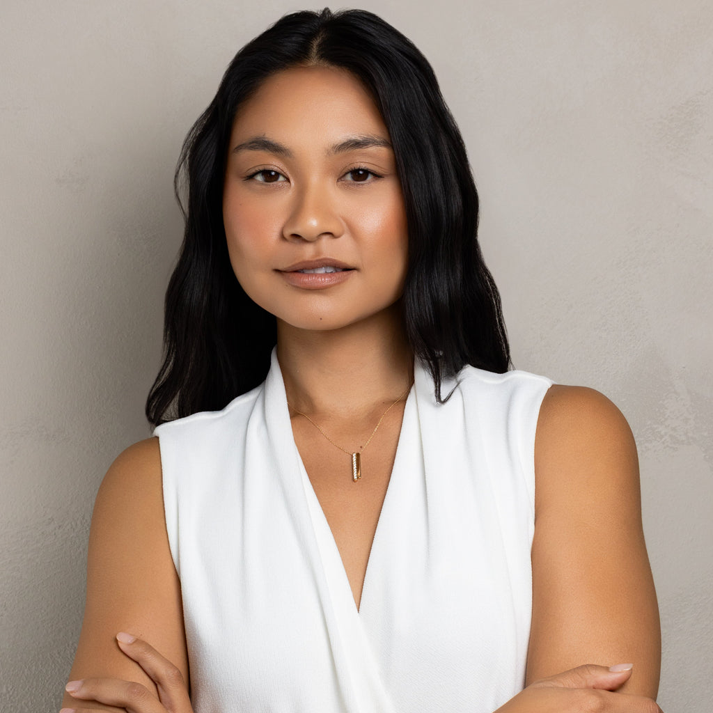 A woman with long dark hair wearing a white sleeveless wrap-style top, standing with her arms crossed against a plain textured beige wall.