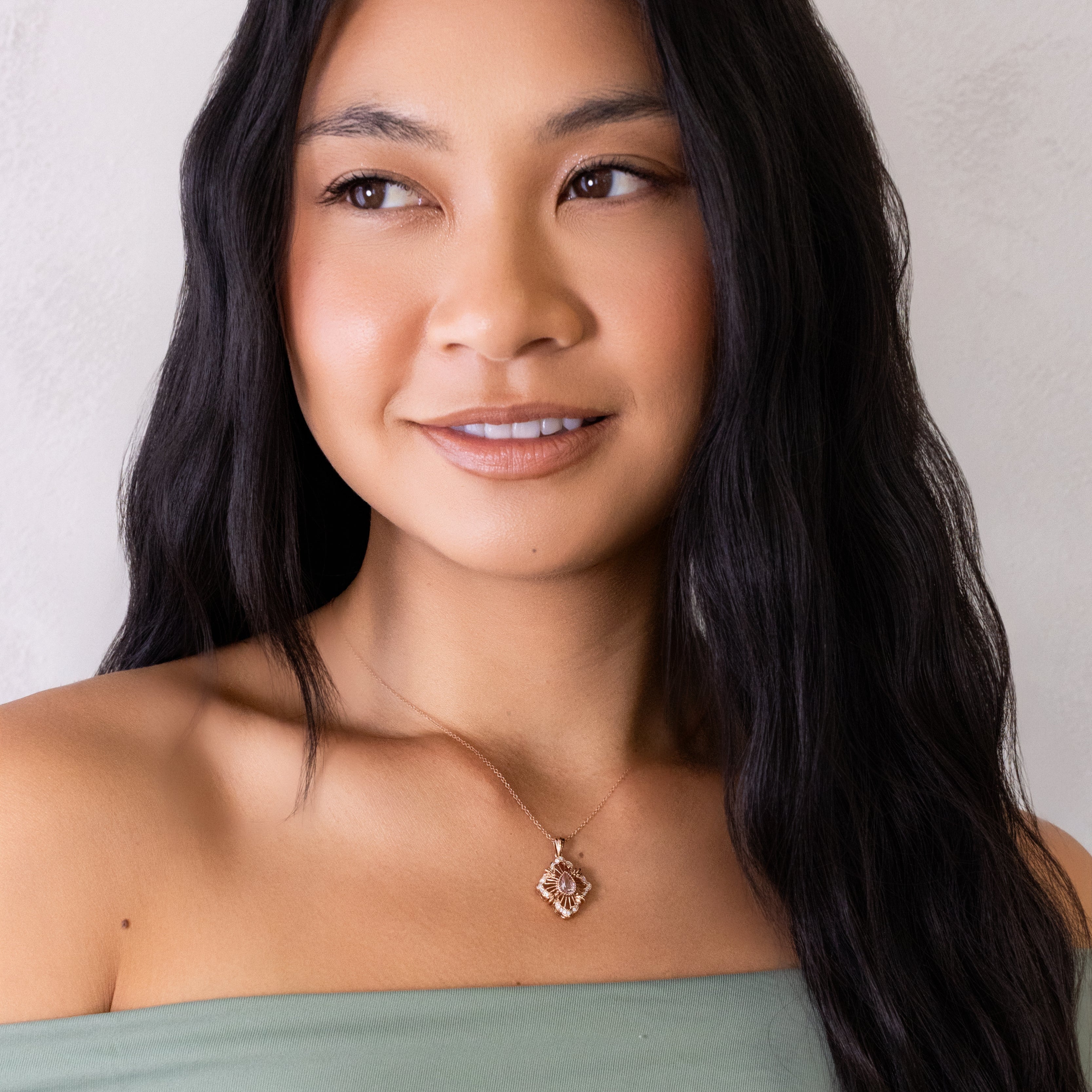 A woman with long, dark wavy hair wearing a sage green off-the-shoulder top and a delicate gold pendant necklace with a geometric diamond design, set against a neutral light gray wall.