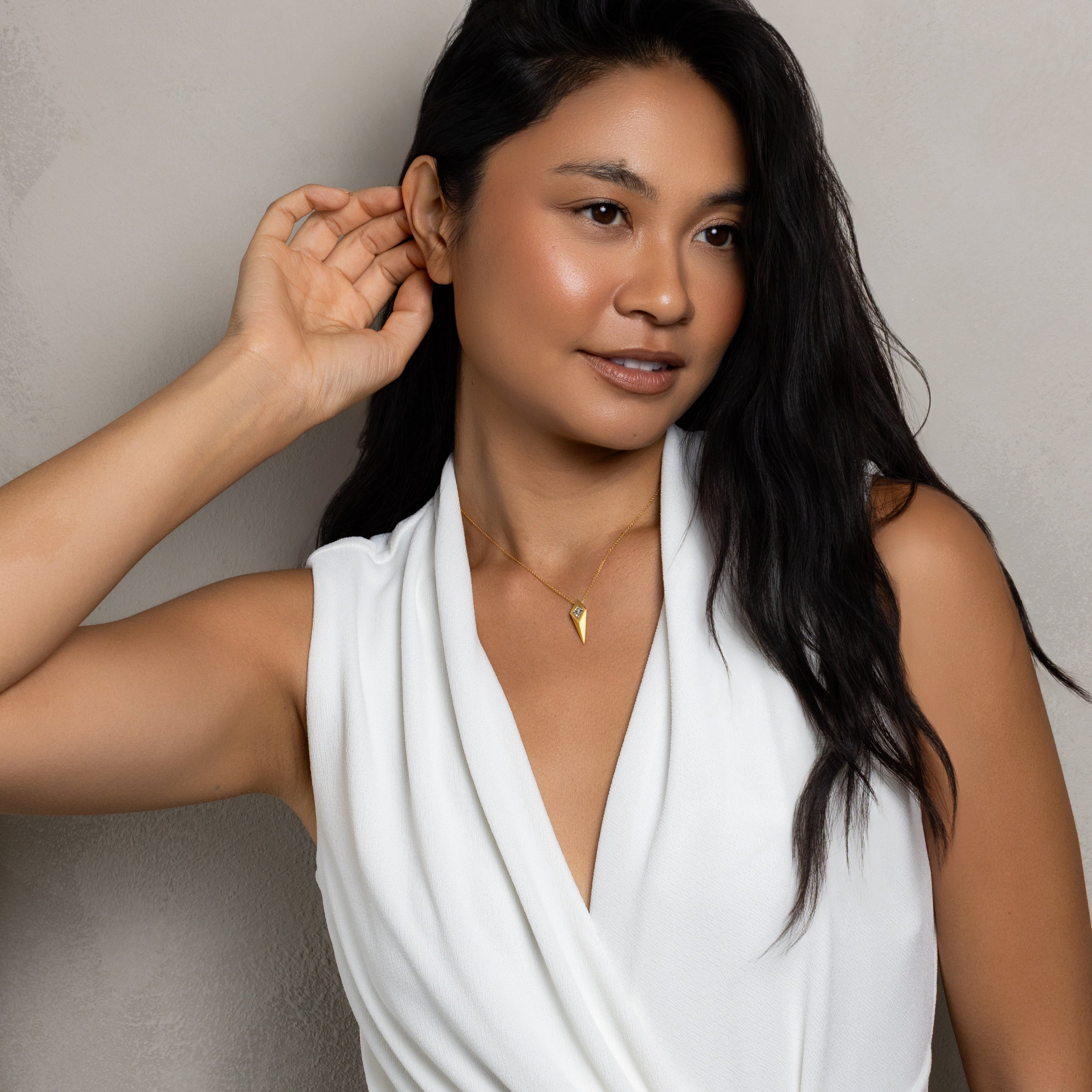 A woman with long dark hair wearing a white draped sleeveless top and a small gold necklace, posing against a textured light gray wall.