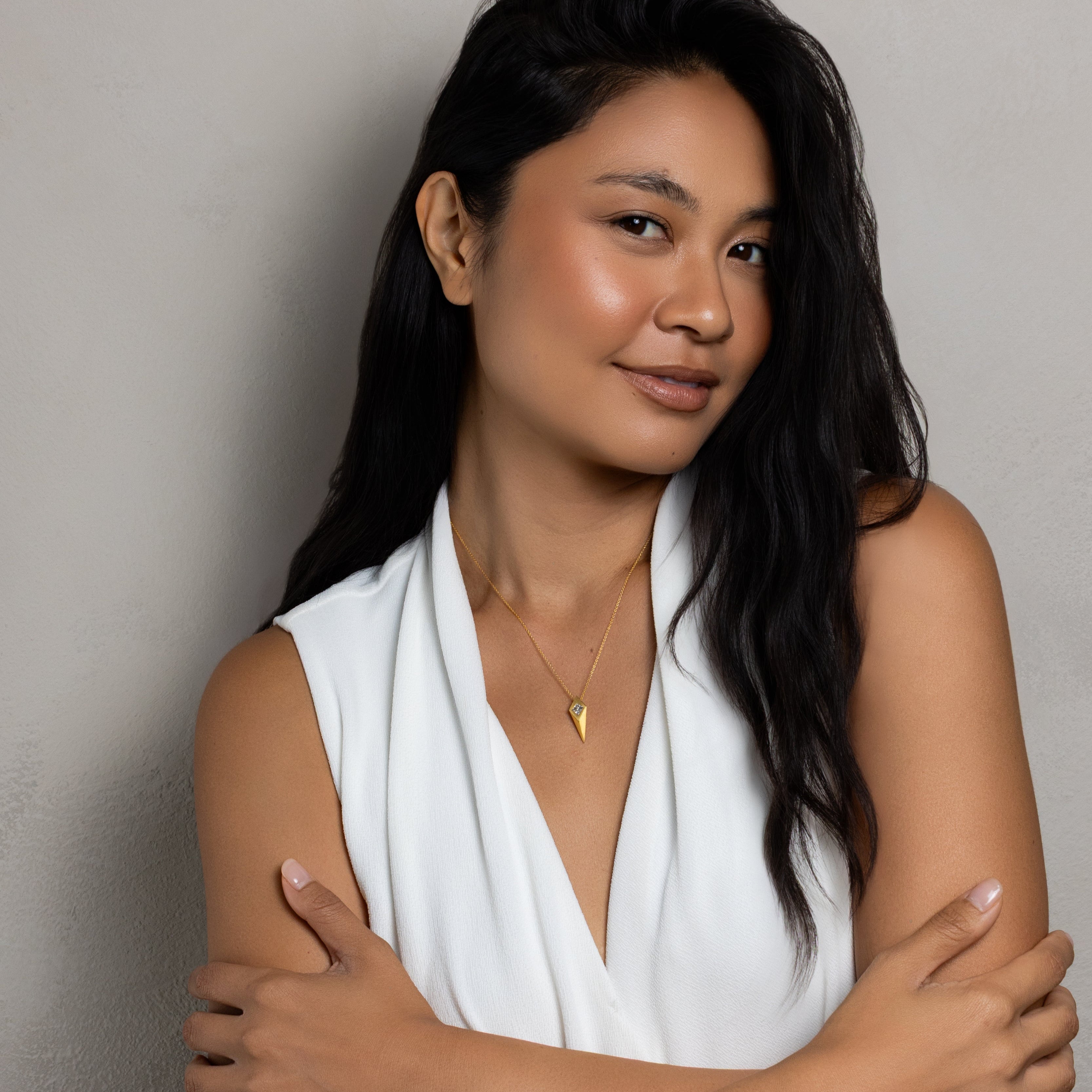A woman with long dark hair wearing a white sleeveless draped top and a gold necklace, posing with her arms crossed against a neutral gray wall.