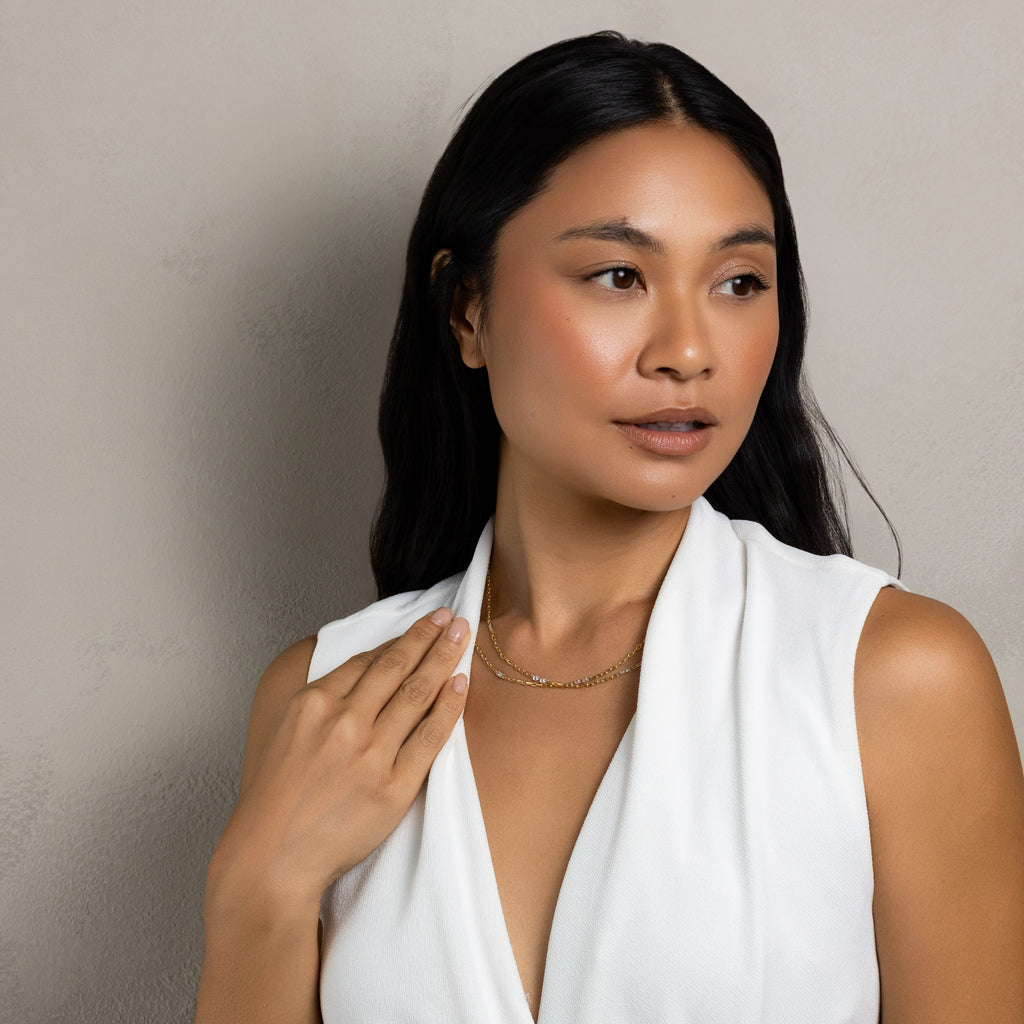 A woman with long dark hair wearing a white sleeveless wrap-style dress, posing against a textured light gray wall.