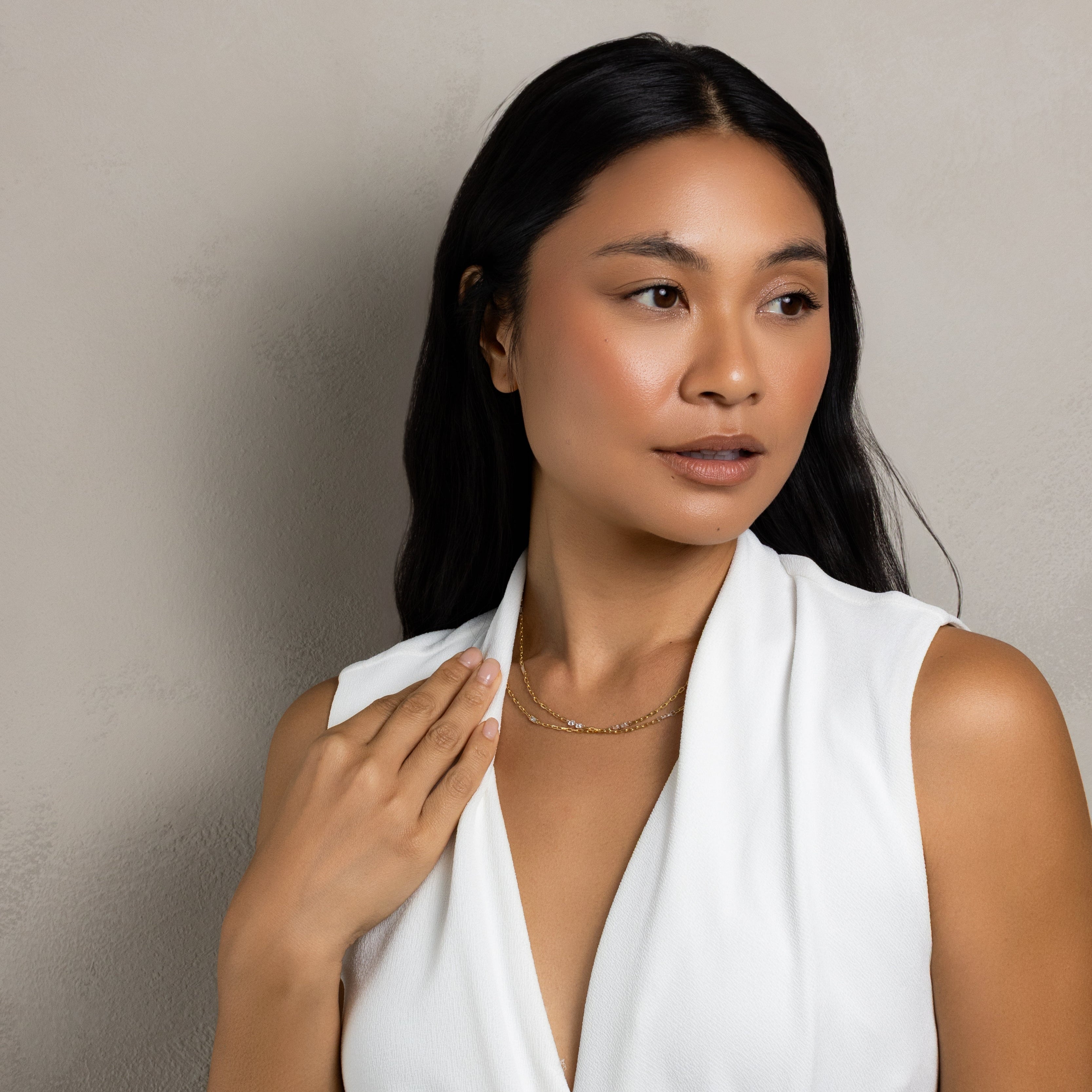 A woman with long dark hair wearing a white sleeveless wrap-style dress, posing against a textured light gray wall.