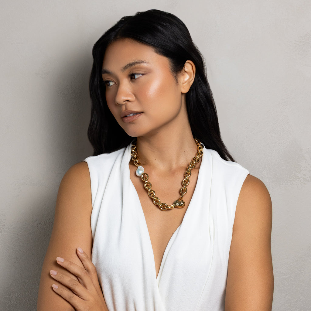 A woman with long black hair wearing a white sleeveless top and a chunky gold chain necklace, standing with her arms crossed against a textured gray wall.