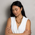 A woman with long black hair wearing a white sleeveless top and a chunky gold chain necklace, standing with her arms crossed against a textured gray wall.