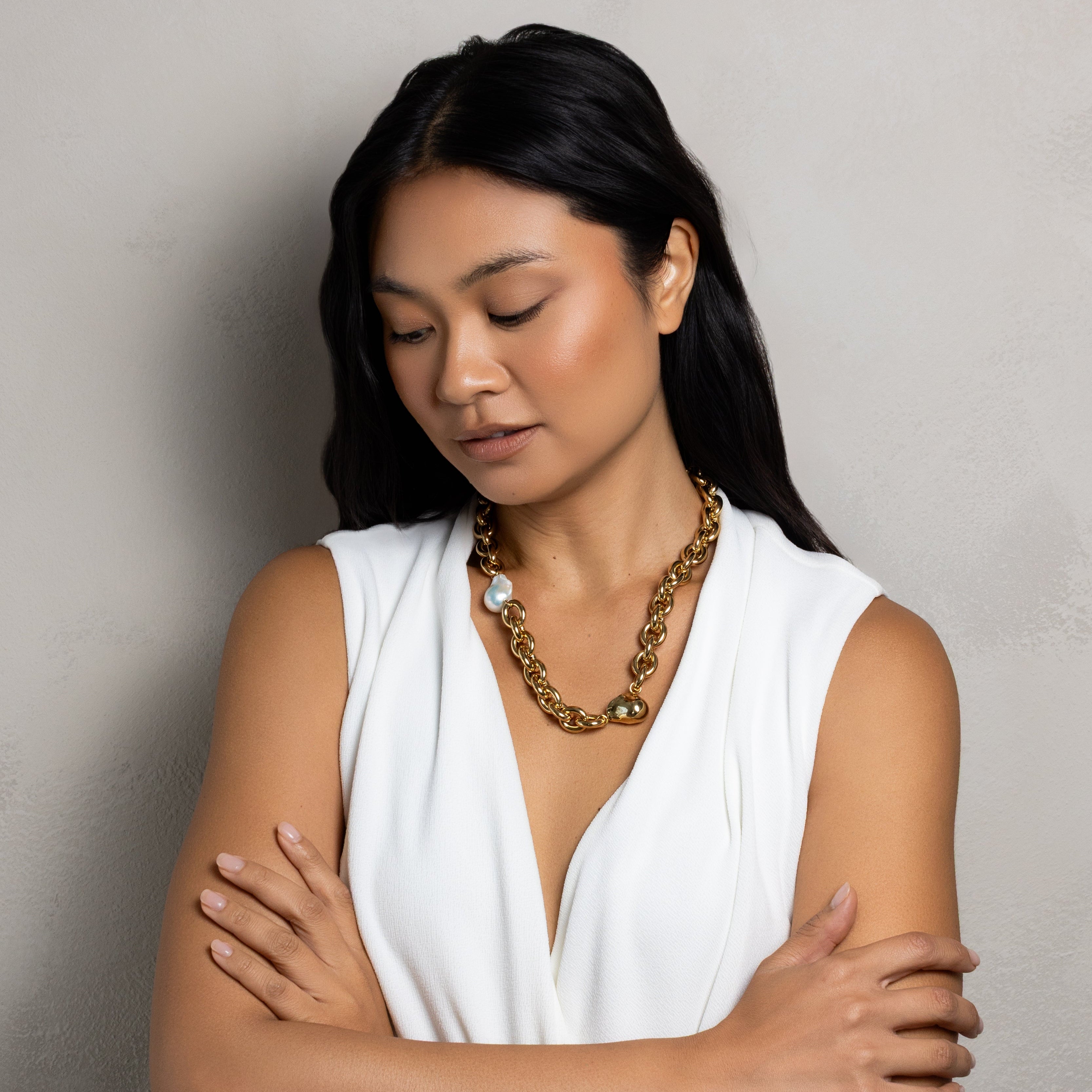 A woman with long black hair wearing a white sleeveless top and a chunky gold chain necklace, standing with her arms crossed against a textured gray wall.