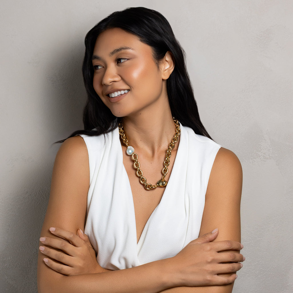 A woman with long black hair wearing a white sleeveless top and a chunky gold chain necklace, standing with her arms crossed against a textured gray wall.