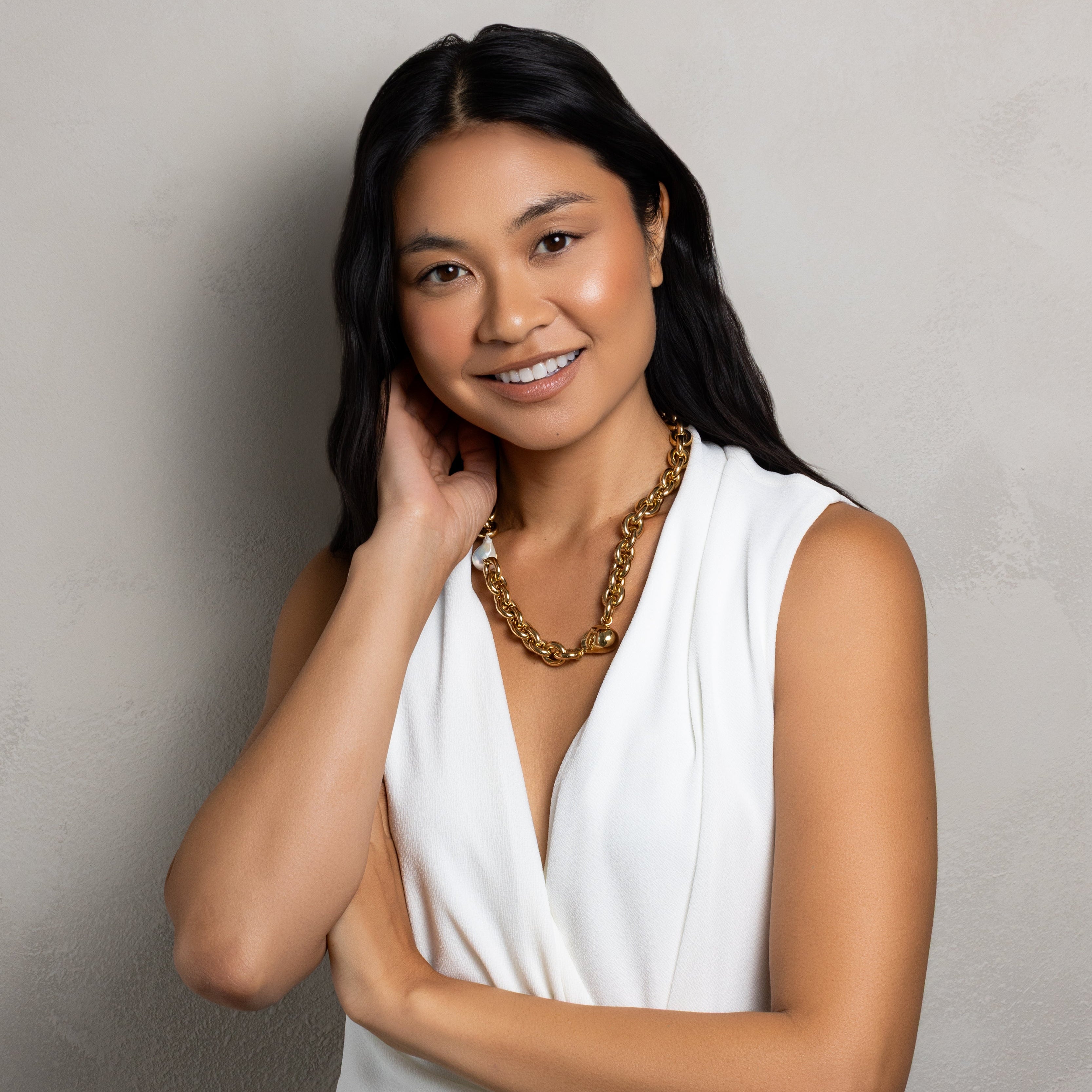 A smiling woman with long dark hair wears a white sleeveless wrap-style dress and a chunky gold chain necklace, posing against a textured light gray wall.
