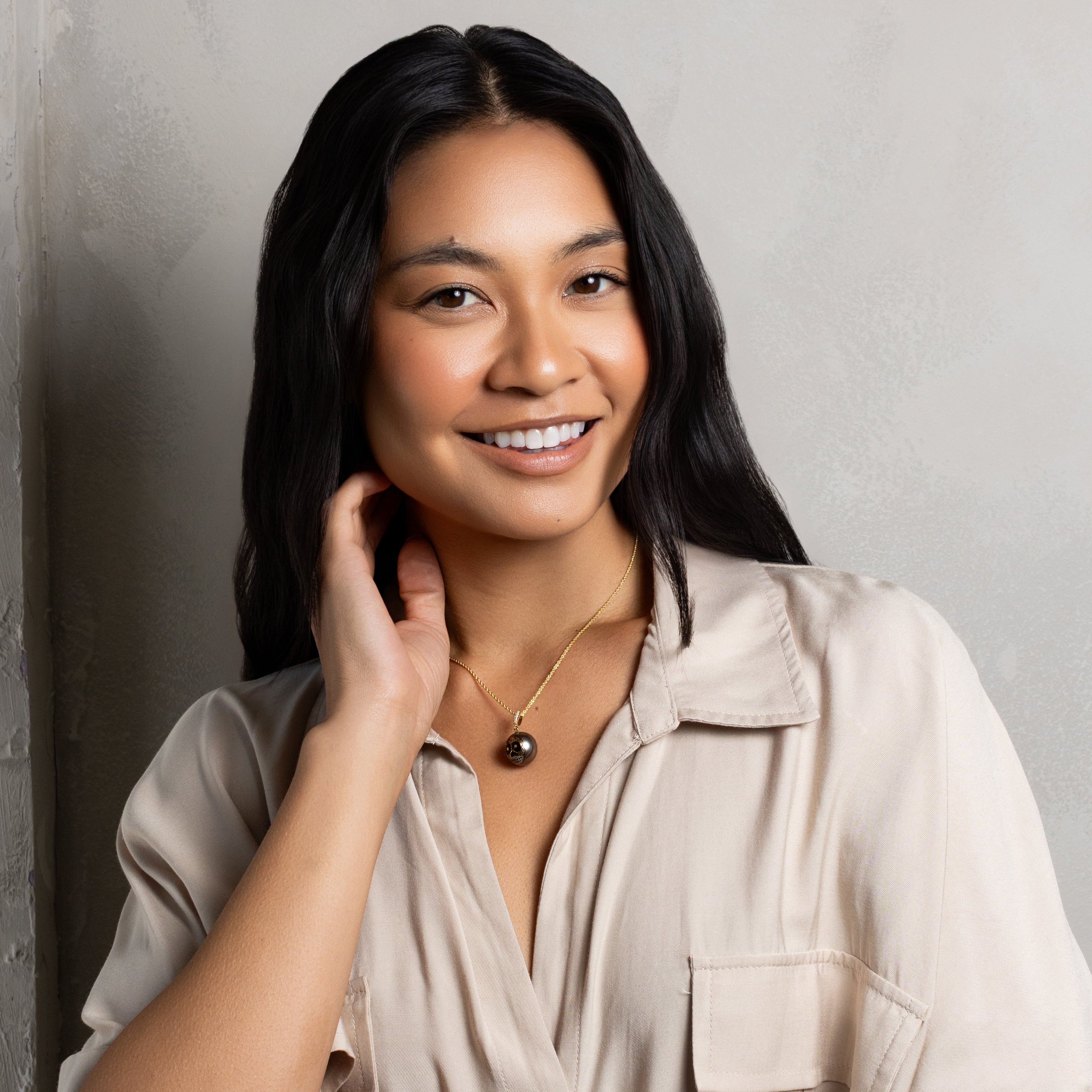 A smiling woman with long black hair is shown from the chest up against a textured light gray wall. She wears a light beige short-sleeve button-up shirt and a delicate gold necklace with a dark round pendant.