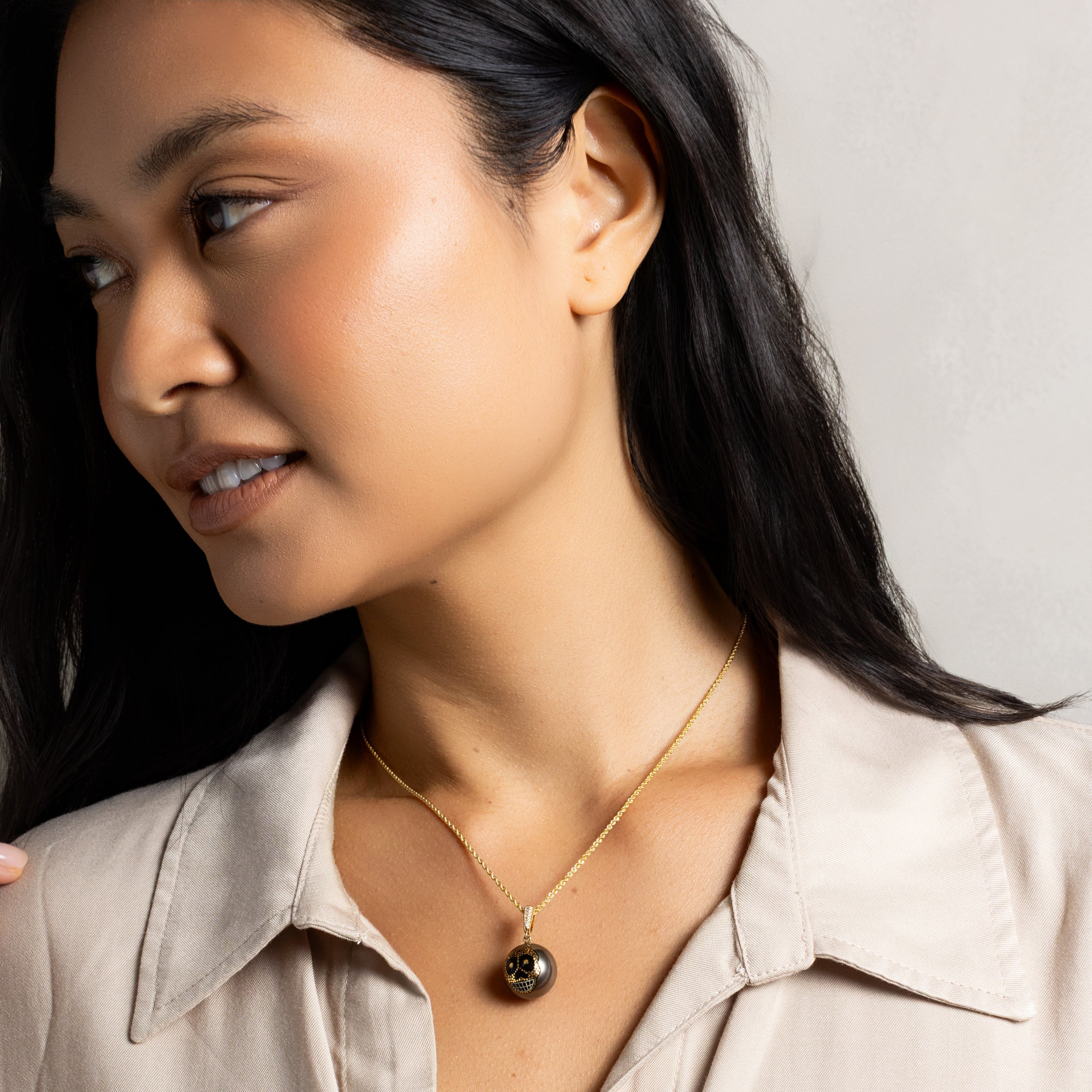 A close-up of a woman wearing a delicate gold chain necklace with a dark, polished round pendant, paired with a light beige button-up shirt.