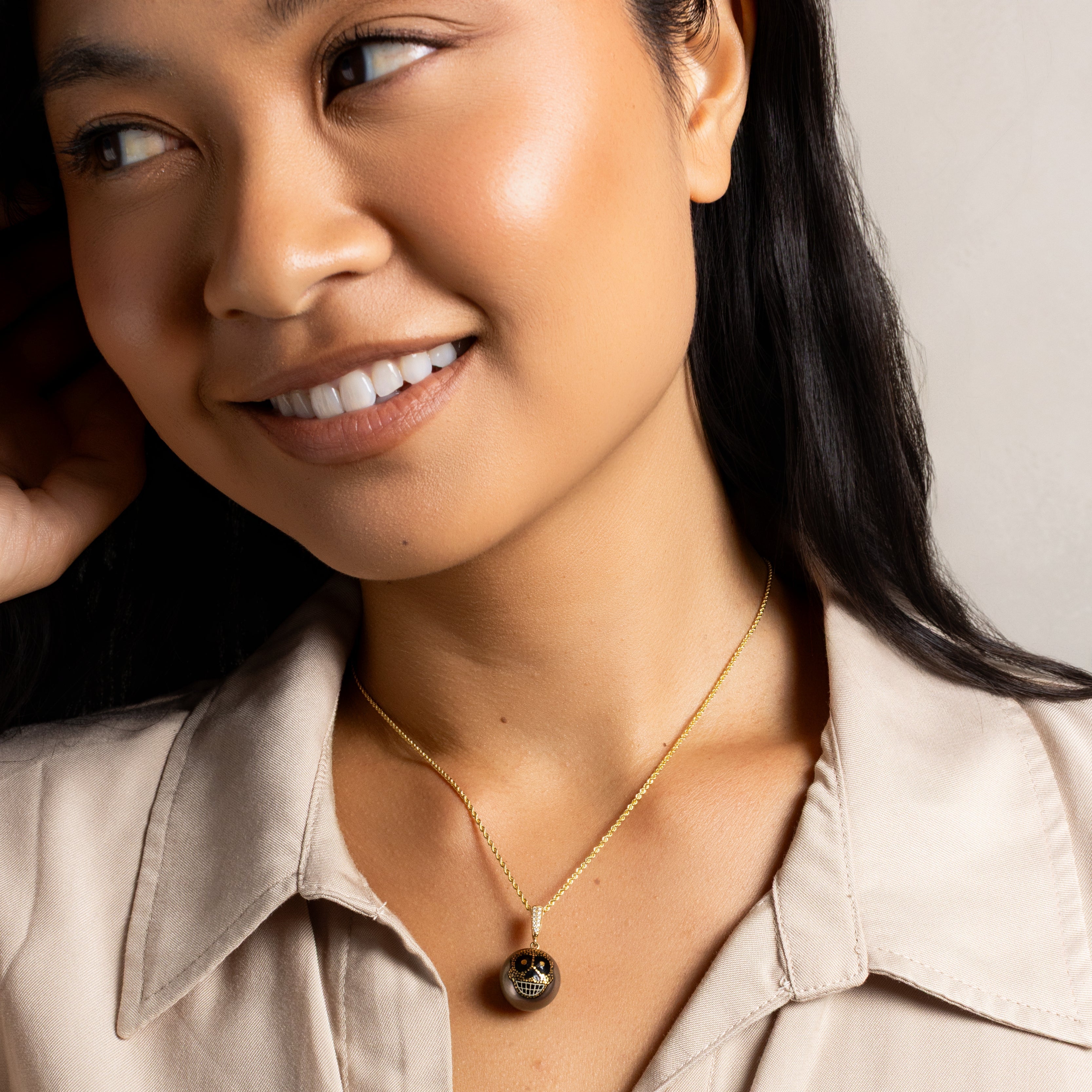A close-up of a woman wearing a delicate gold chain necklace with a dark, round, textured pendant, paired with a beige collared shirt.