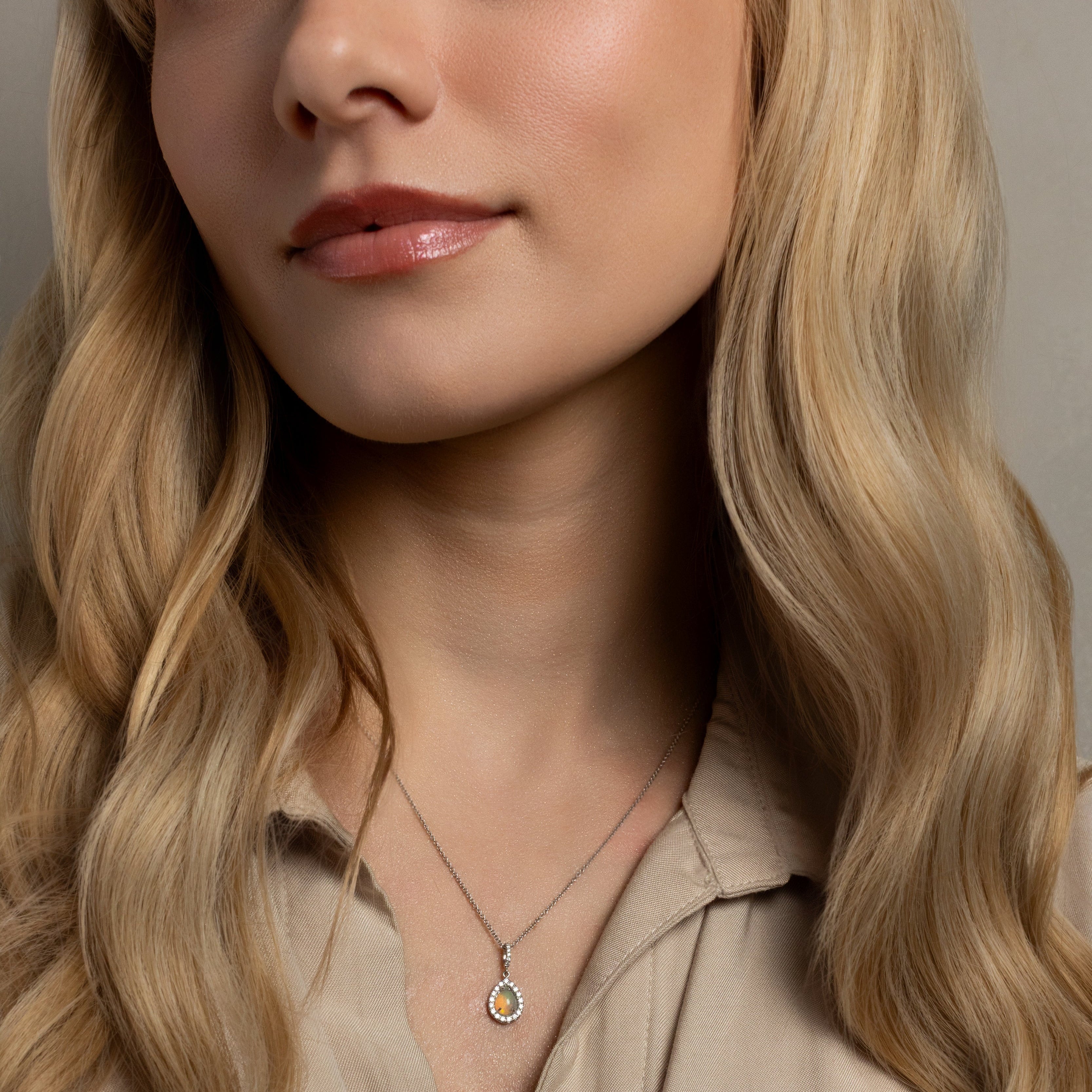 A close-up of a blonde woman wearing a delicate silver pendant necklace with a small circular blue stone, paired with a beige collared shirt.