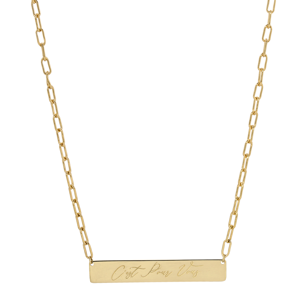 A gold bar necklace with a rectangular pendant engraved with the words 'Just Like That' in cursive script, suspended from a paperclip chain on a white background.