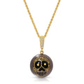 A gold chain necklace with a round pendant featuring a black and gold sugar skull design on a smoky brown background, set against a plain white background.